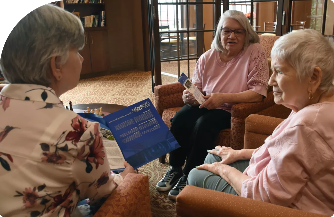 windsor-house-facility-conversation Elderly women smiling while playing cards with a caring nurse at Windsor House senior care community.
